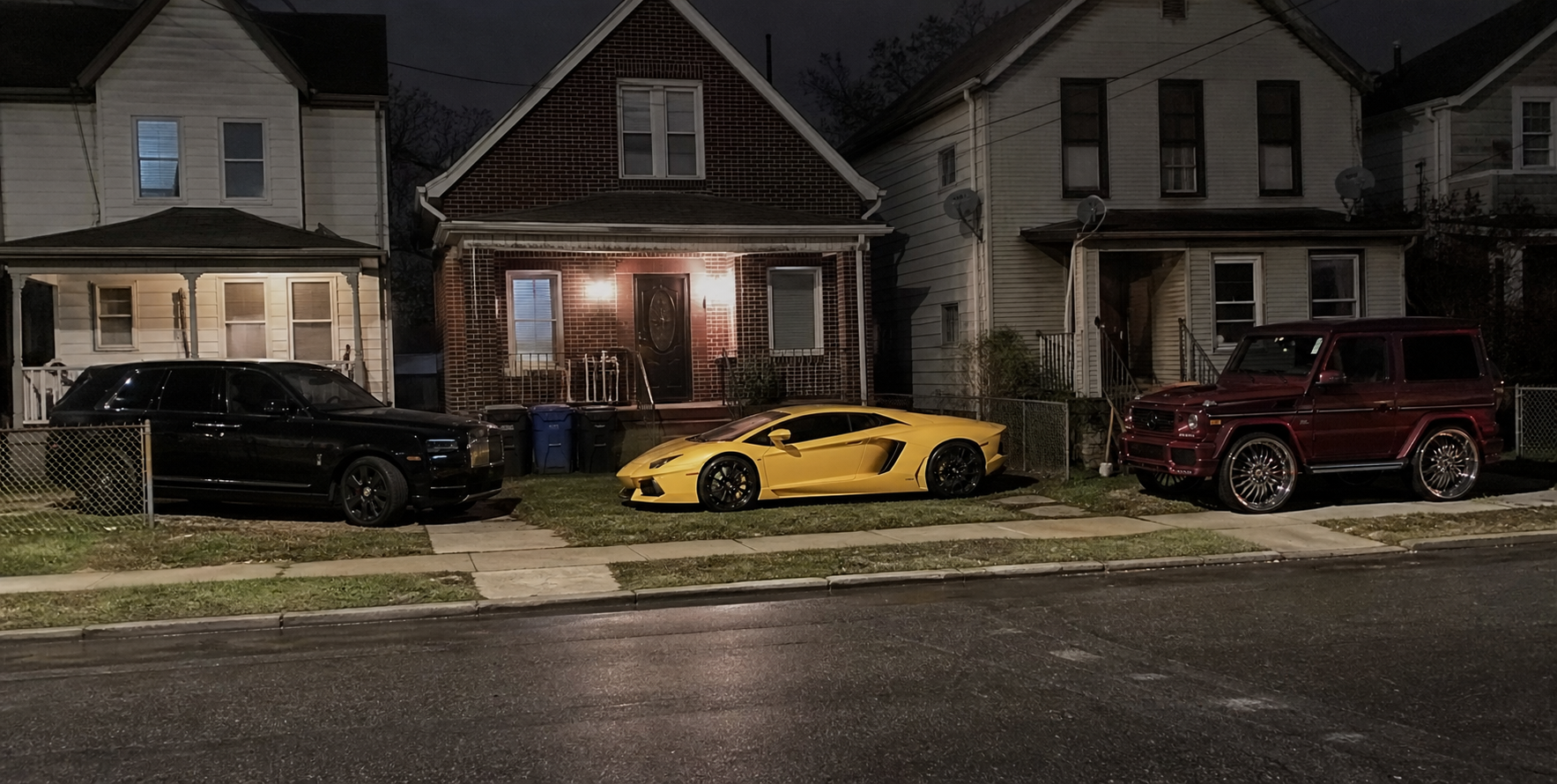 Three luxury vehicles outside modest neighboring houses at night